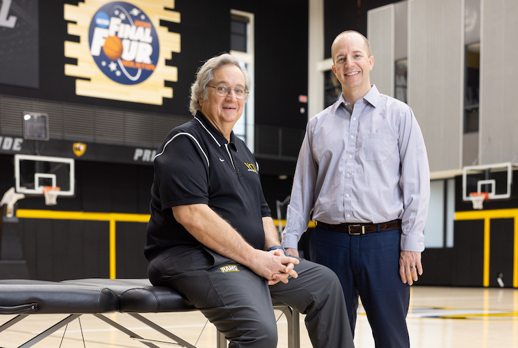 The two men are on a VCU basketball court, Thomas is sitting down while Seth is standing. Both are smiling towards the camera.