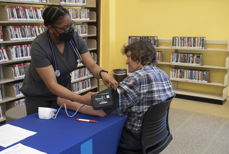 Student measures the blood pressure of a seated man. They are surrounded by bookshelves.