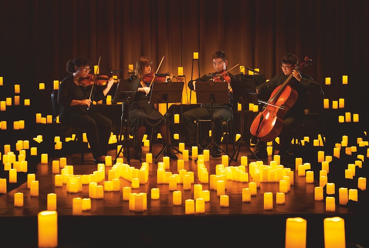 Four students playing stringed instruments on a stage with hundreds of electric candles around them. 