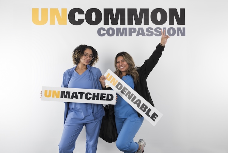 Two female VCU Health team members hold up signs and smile for a photo. One team member is raising her hand with a peace sign.