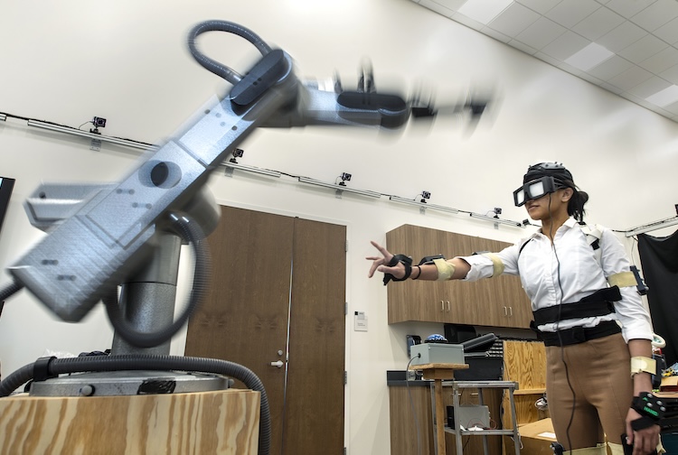 Female student, wearing large glasses over her eyes, reaches for a robot. She is demonstrating the physical component of the research project, testing hand-eye coordination.  