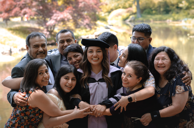 Woman featured throughout this story stands in the middle of a big group huge with family members. All of them are smiling, proud of her for graduating.