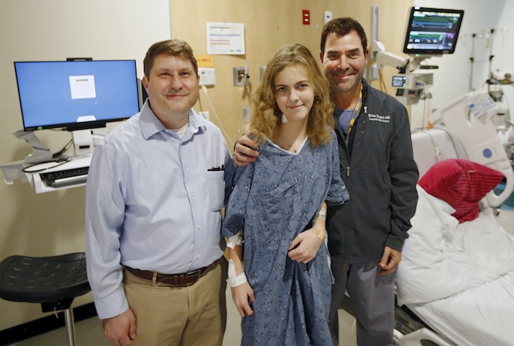 A father, his daughter and a doctor stand together in a hospital room smiling.