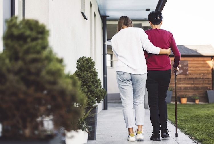 A caregiver helps an older woman walk in front of her house