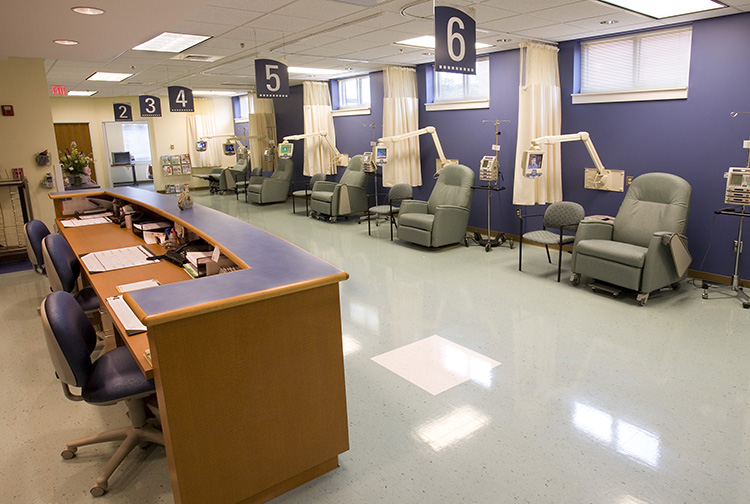 A room with a nursing station and six bays with green chairs and medical equipment in each.