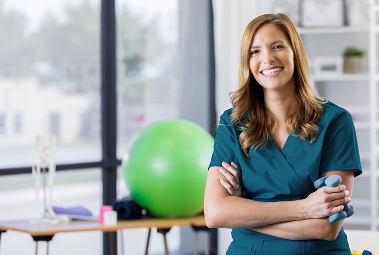 A physical therapist wearing teal scrubs smiles with a bright green exercise ball in the background.