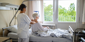 Woman sitting on a hospital bed looking out the window. She is having her neck massaged by a healthcare provider.