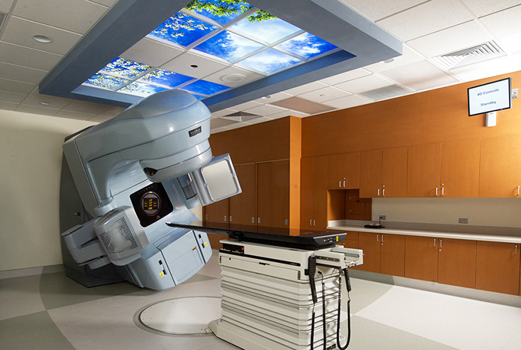 Equipment that delivers radiation for cancer patients in a room with a lit ceiling resembling trees and sky.