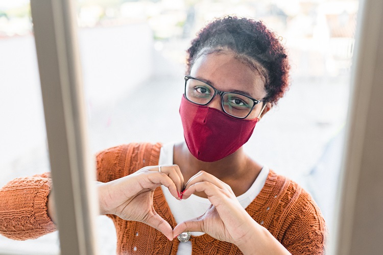 Woman wearing a mask and making a heart shape with her hands through a window