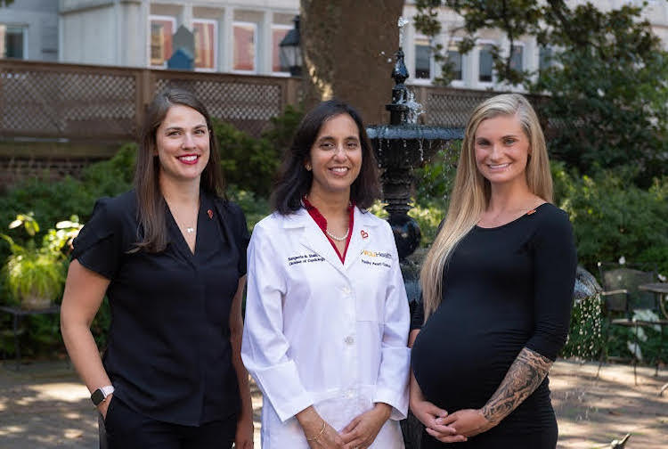 Three women stand outside smiling. Two are wearing black outfits while the woman in the middle, Dr. Shah, is wearing her white doctor's coat.